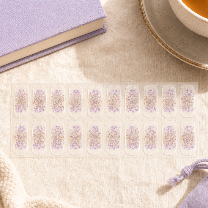 A sheet of 15 clear press-on nails with lavender floral and gold glitter designs, arranged on a beige linen surface next to a purple book and a cup of tea.